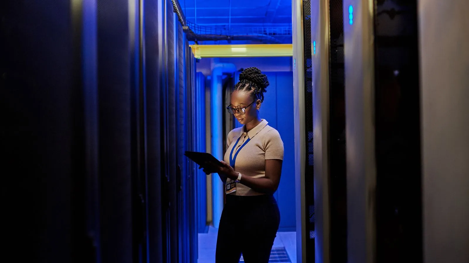 A woman holding a tablet in a blue-lit data center aisle lined with server racks.