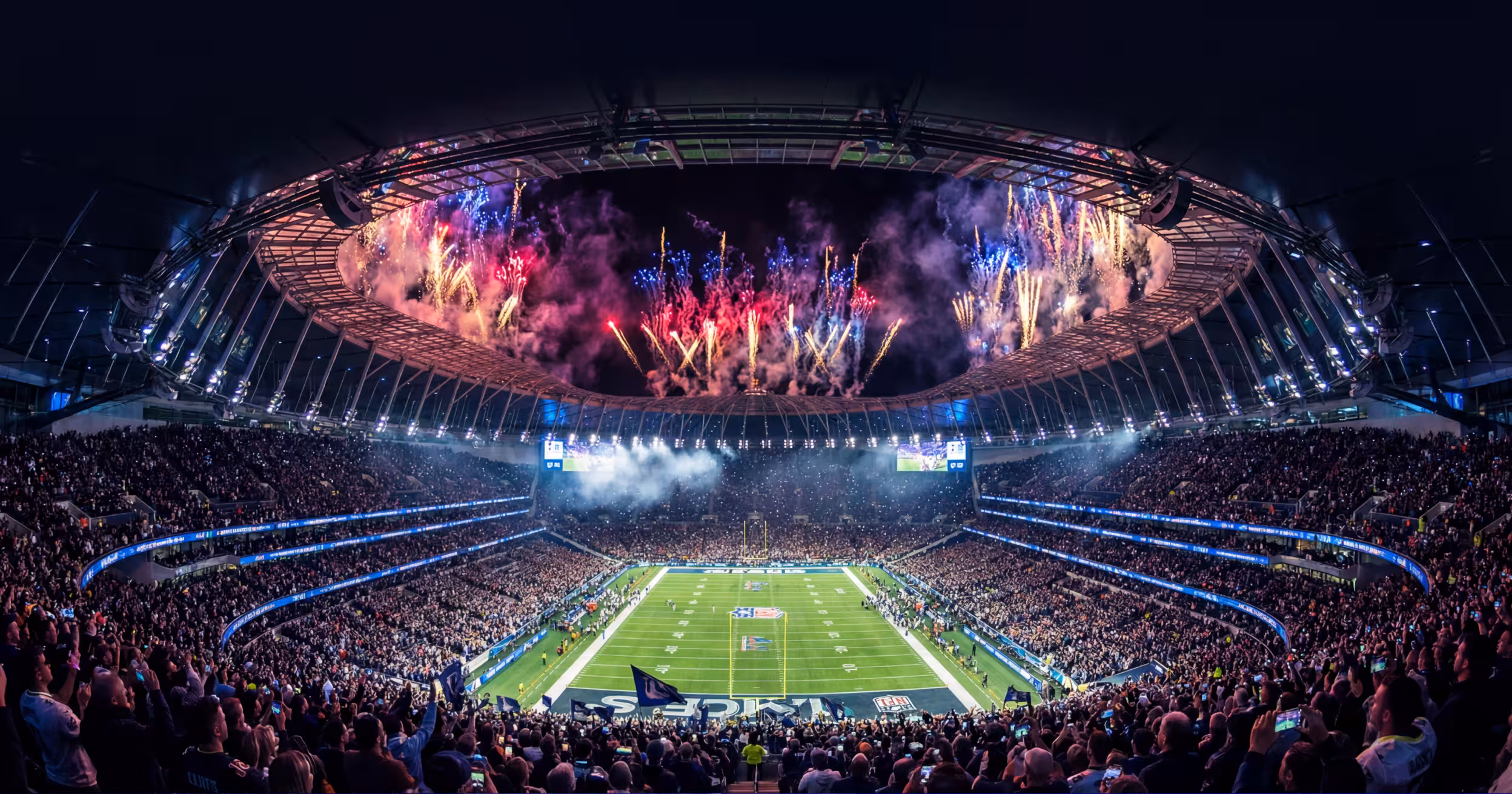 A packed stadium at night, fireworks lighting up the sky above the floodlit pitch.
