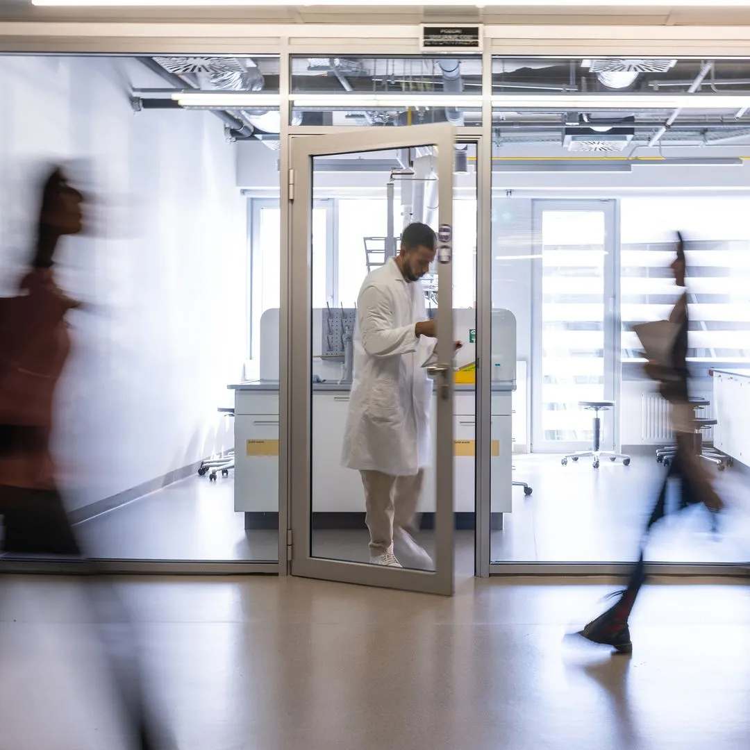 A man in a lab coat walking through a glass door in a research lab, others passing in motion blur