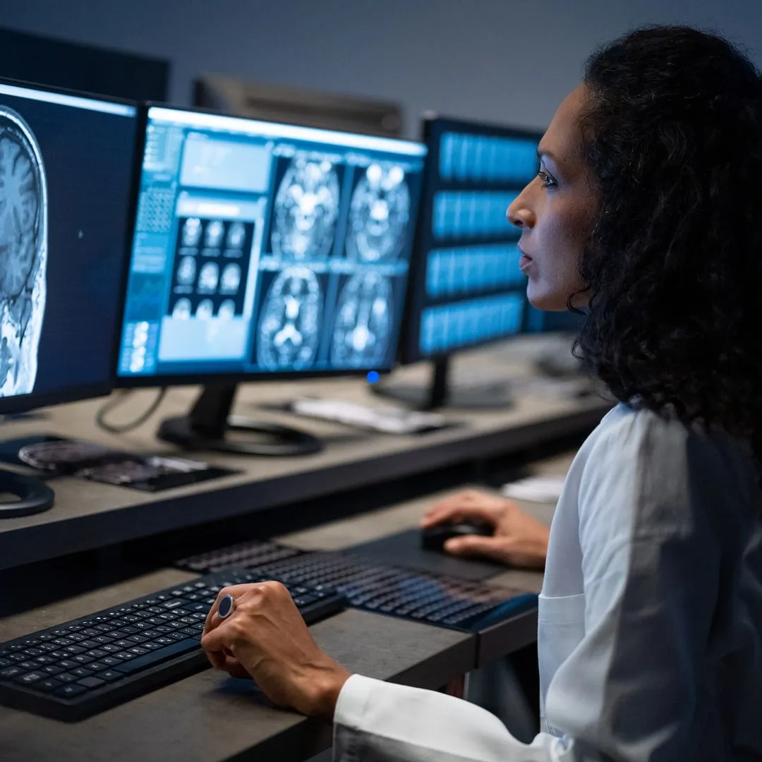 A medical professional viewing brain-scan images across multiple monitors in a dim reading room