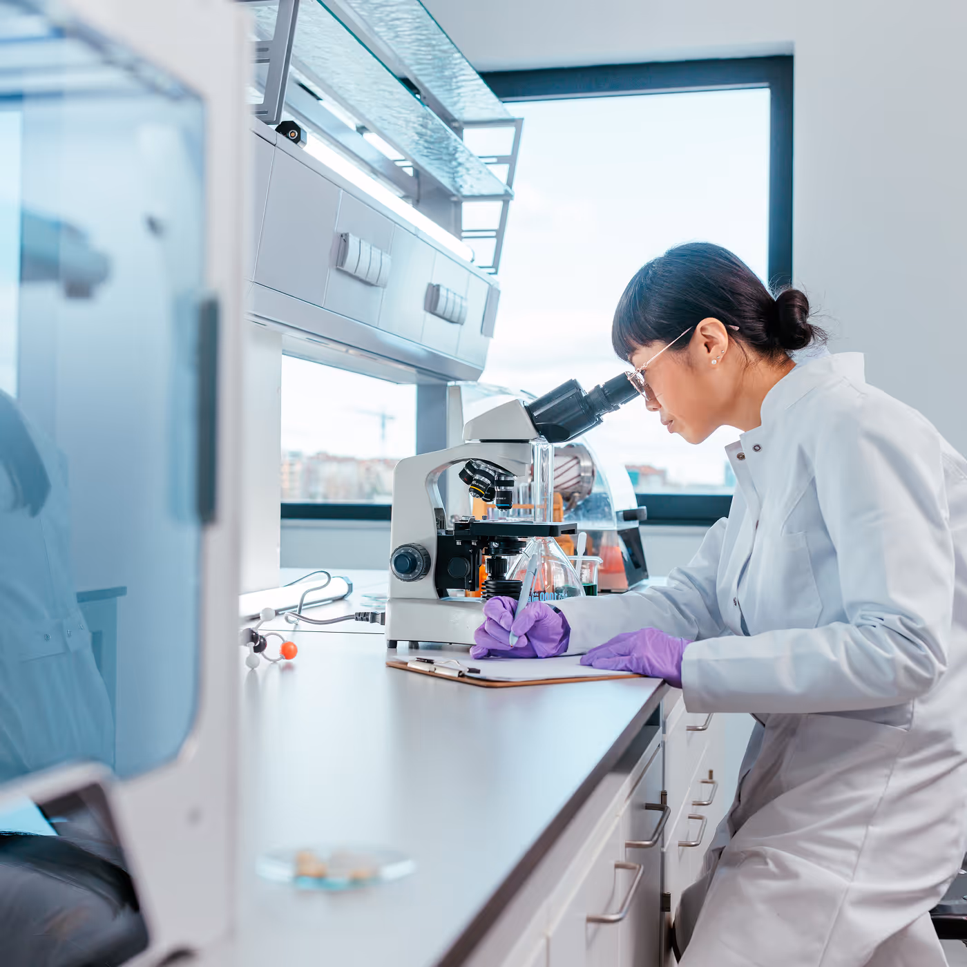 A scientist in a white coat and gloves looking through a microscope at a lab bench by a window