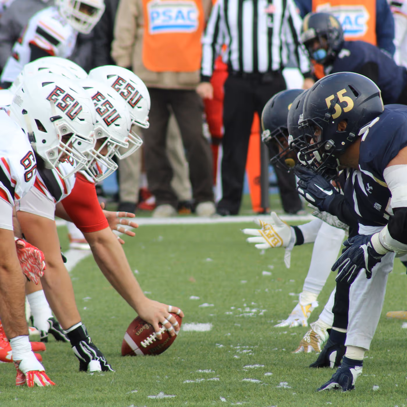 College football players lined up at the line of scrimmage just before the snap, with a referee in the background