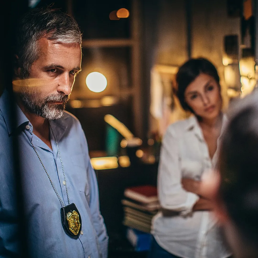 A law enforcement officer wearing a badge on a neck chain, in conversation indoors