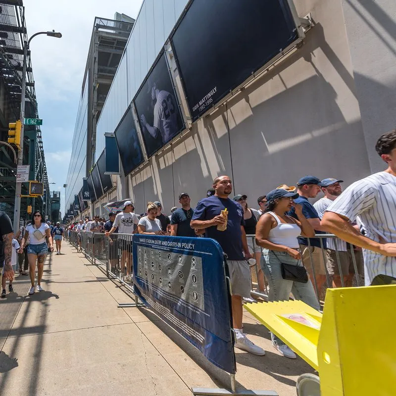 Fans waiting in line behind barriers along a sidewalk outside a sports stadium on a sunny day