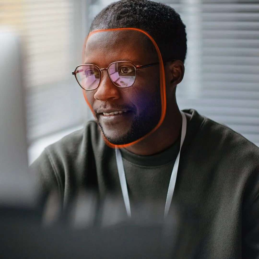 Man wearing glasses at a desk, his face outlined by an orange facial-recognition scan overlay