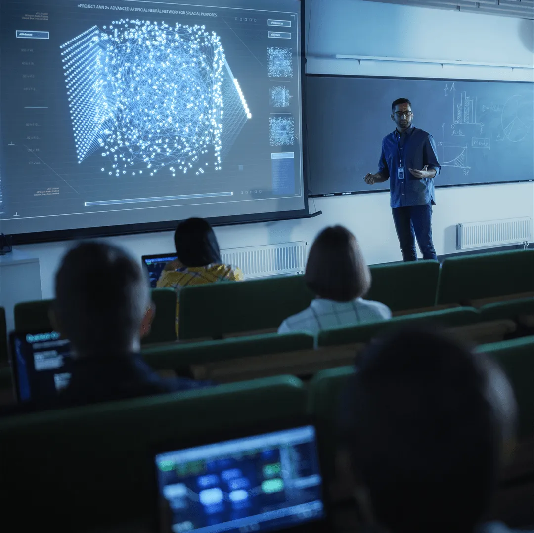 A lecturer presenting to students in a tiered hall, with a large screen showing a neural-network visualization