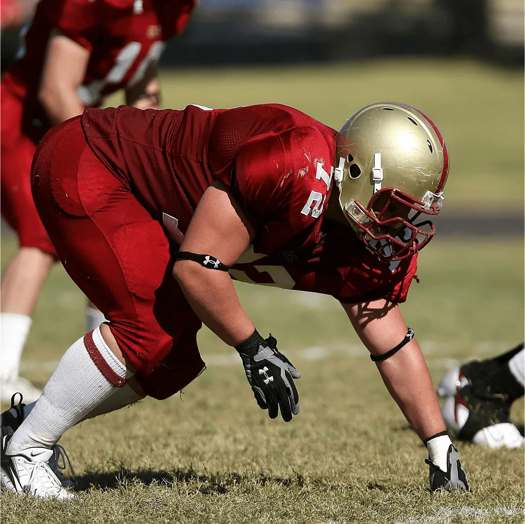 A college football player in a red uniform and gold helmet crouched in a stance on the field