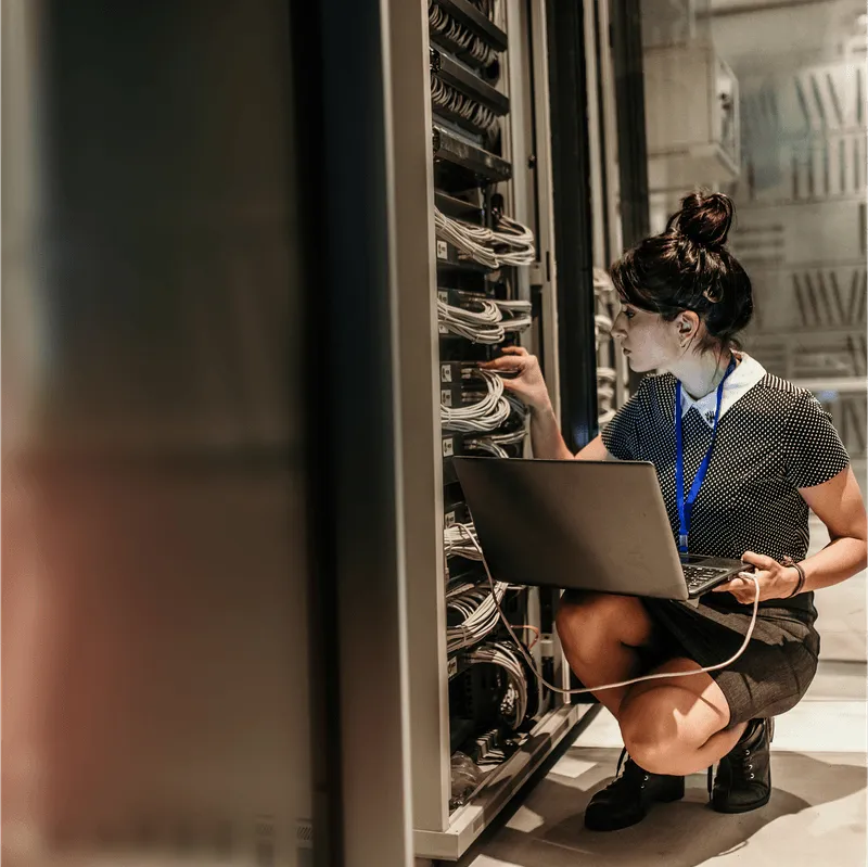 A technician crouching with a laptop while working on cabling inside a data center server rack