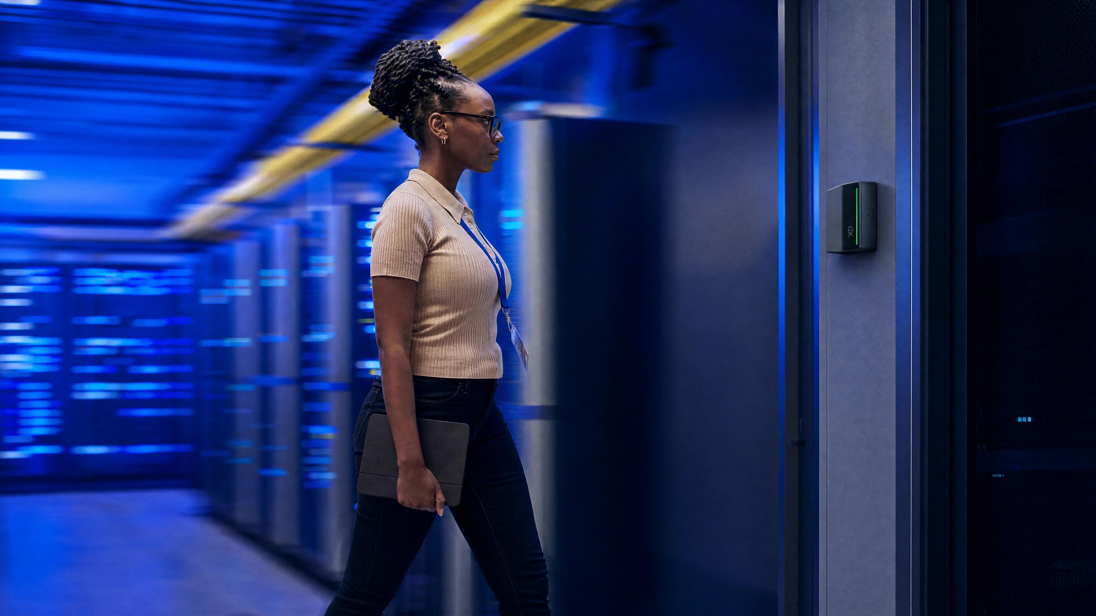 A technician walking toward a data hall door where a Rock X device glows green, server racks lit blue behind her