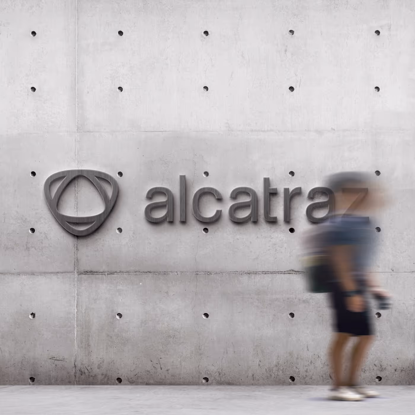 Person walking past a concrete wall carrying the brushed-metal Alcatraz logo and wordmark, captured in motion blur