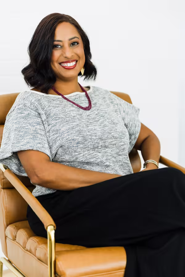 Smiling woman with dark hair sitting in a tan chair wearing a gray top and black pants.