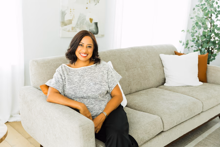 Smiling woman with short dark hair sitting on a light gray sofa in a bright living room.