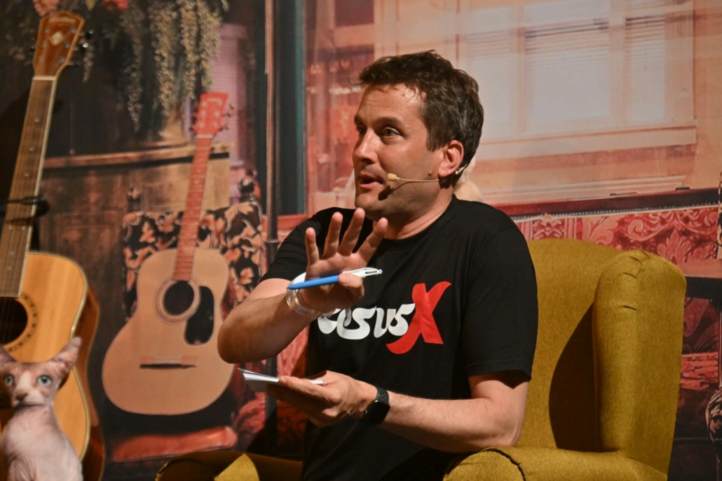 Iain Beardsell seated on stage, wearing a headset microphone and gesturing during a discussion.