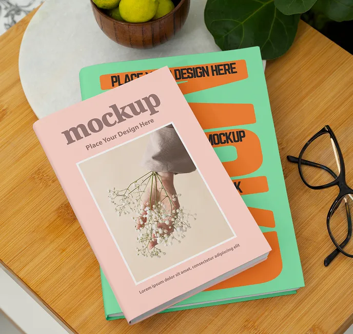 Two books stacked on a wooden table near black framed glasses; the top book has a pink cover with an image of a hand holding white flowers and the text 'mockup Place Your Design Here'.