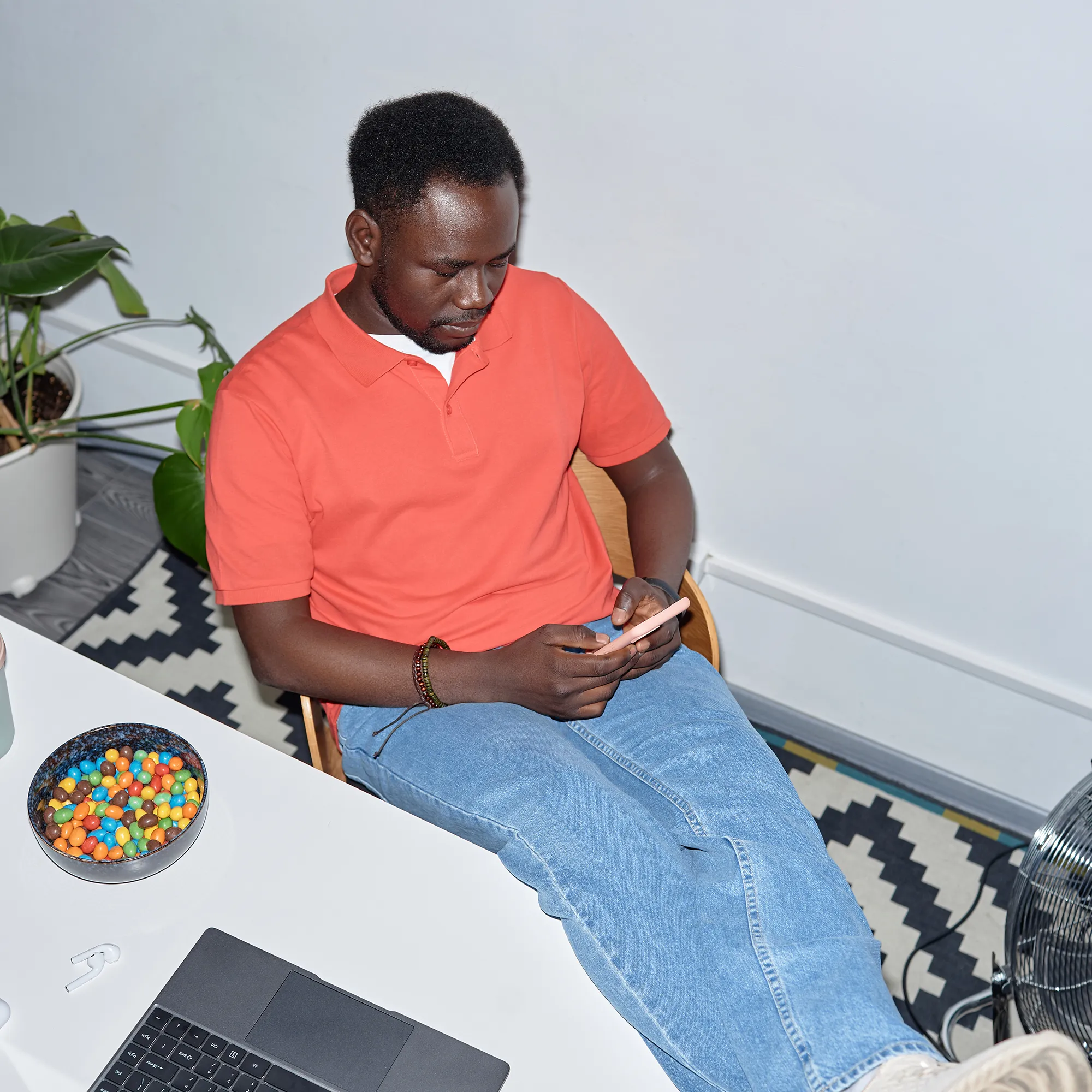 Man in een oranje shirt gebruikt zijn smartphone naast een laptop