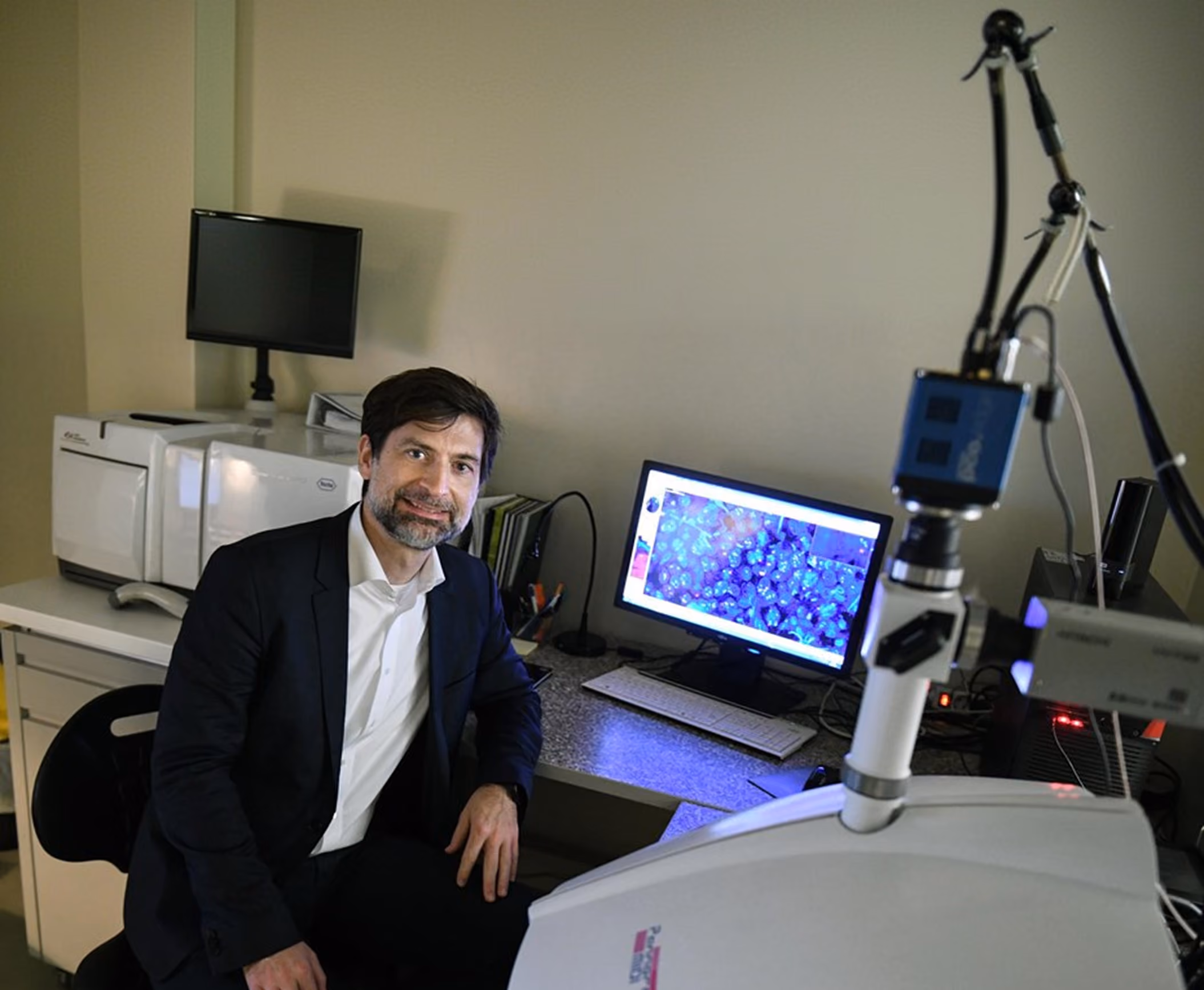 Dr. Peták István sitting in a laboratory beside a microscope with a computer monitor showing a magnified image of blue cells or tissue samples.