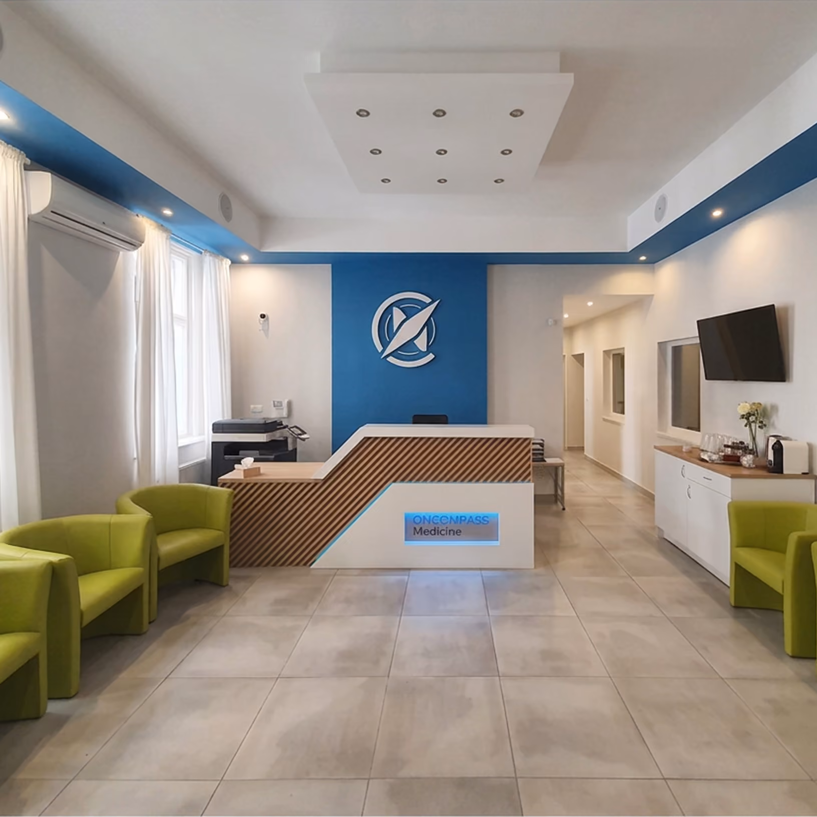 Oncompass Medicine office reception area with a white and wood reception desk labeled Oncompass Medicine, blue accent wall with logo, green chairs, large windows with white curtains, tiled floor, a flat-screen TV on the right wall, and a white air conditioner above the windows.