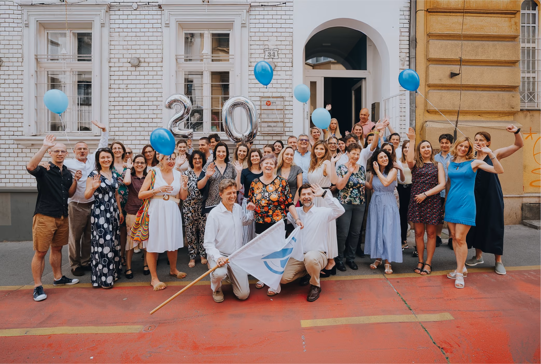 Oncompass team gathered outside a building, smiling and waving at the camera, holding blue balloons and silver balloons shaped as the numbers '2' and '0', celebrating an event with two men kneeling at the front holding a white flag with Oncompass Medicine logo.