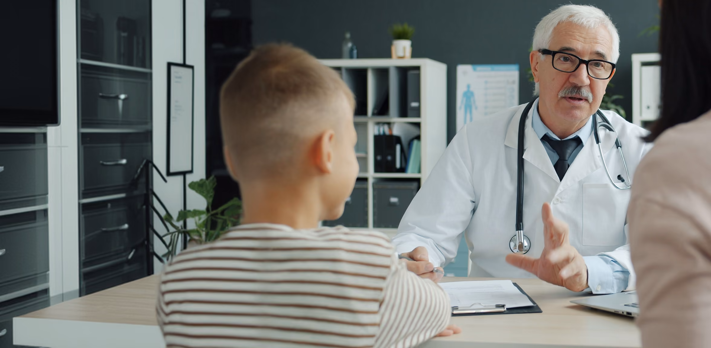 A male doctor with white hair, glasses, and a mustache wearing a white coat and stethoscope talks to a woman and child across a desk in a medical office.