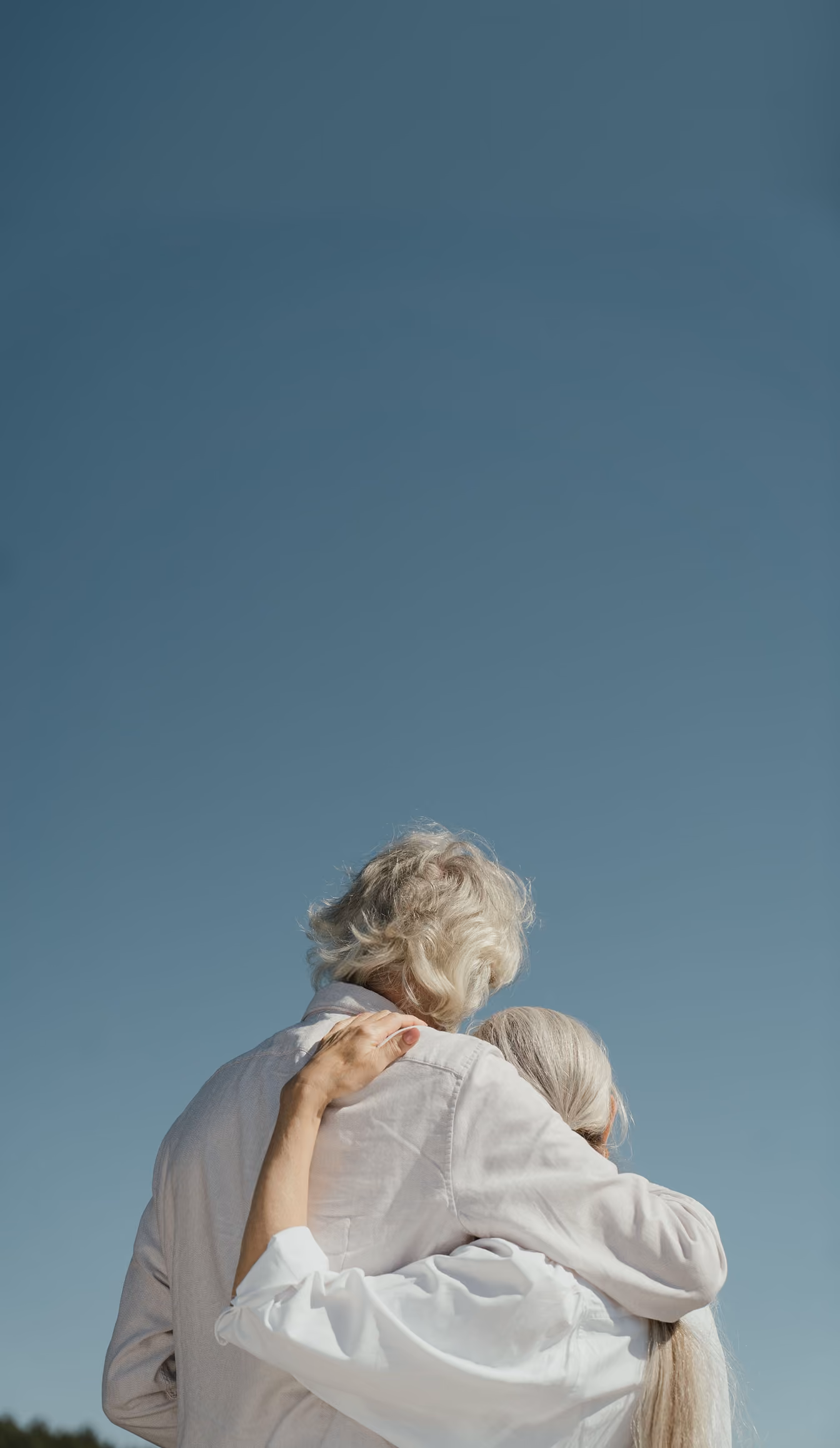 Older couple with gray hair embracing outdoors under clear blue sky, seen from behind.