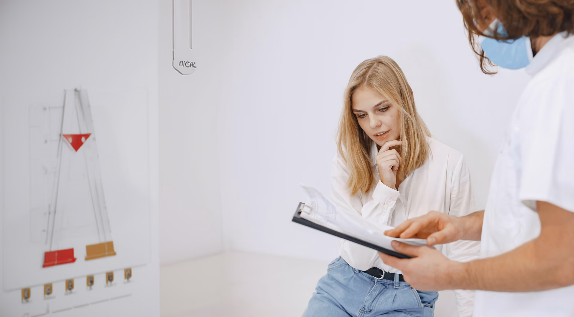 A female patient in a white blouse and blue jeans seated, looking thoughtfully at a clipboard held by a male healthcare professional wearing a white coat and blue face mask.