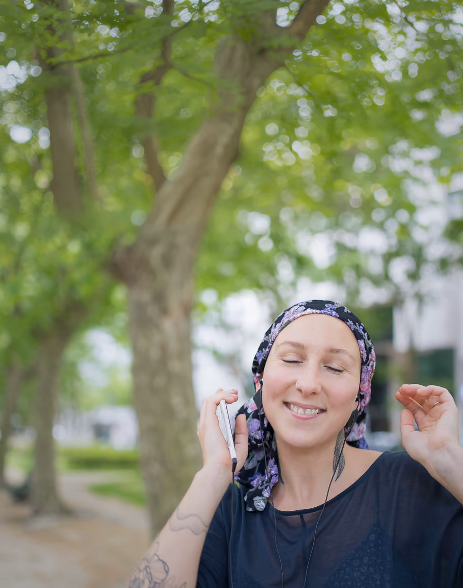 Smiling woman with a floral headscarf and tattoos on her arm listens to music with earphones outdoors in a park, holding a phone and raising one hand.