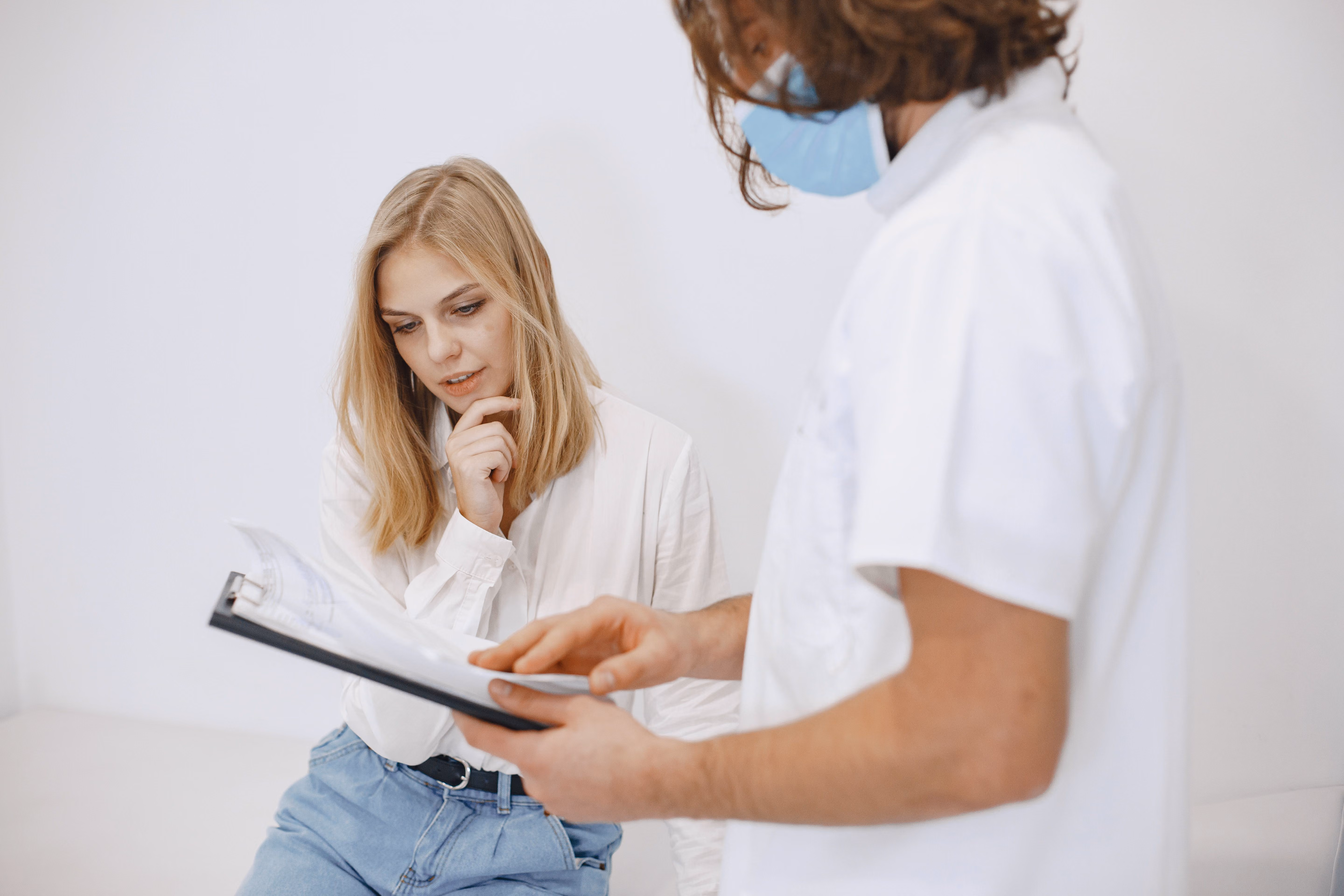 A female patient in a white blouse and blue jeans seated, looking thoughtfully at a clipboard held by a male healthcare professional wearing a white coat and blue face mask.