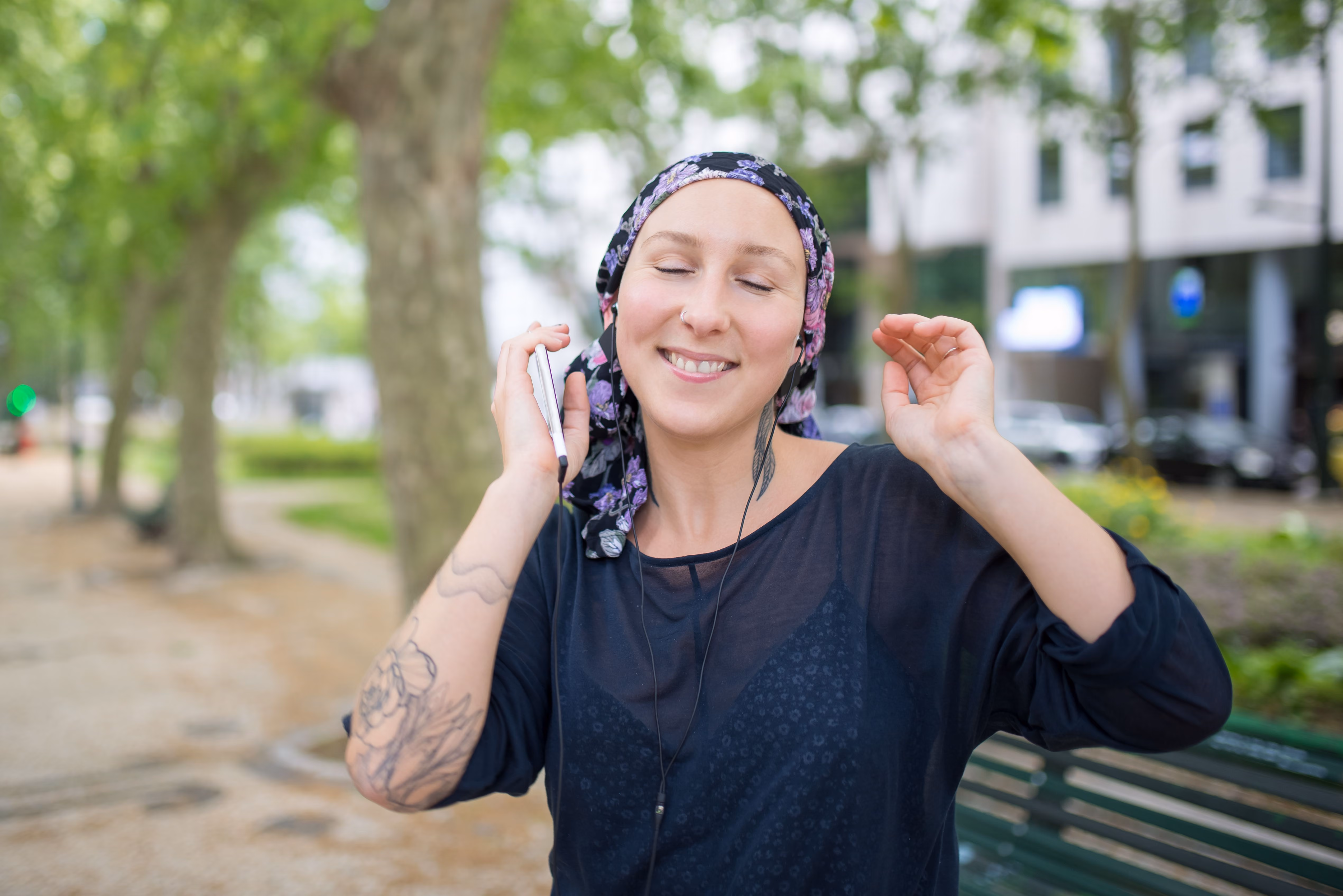 Smiling woman with a floral headscarf and tattoos on her arm listens to music with earphones outdoors in a park, holding a phone and raising one hand.