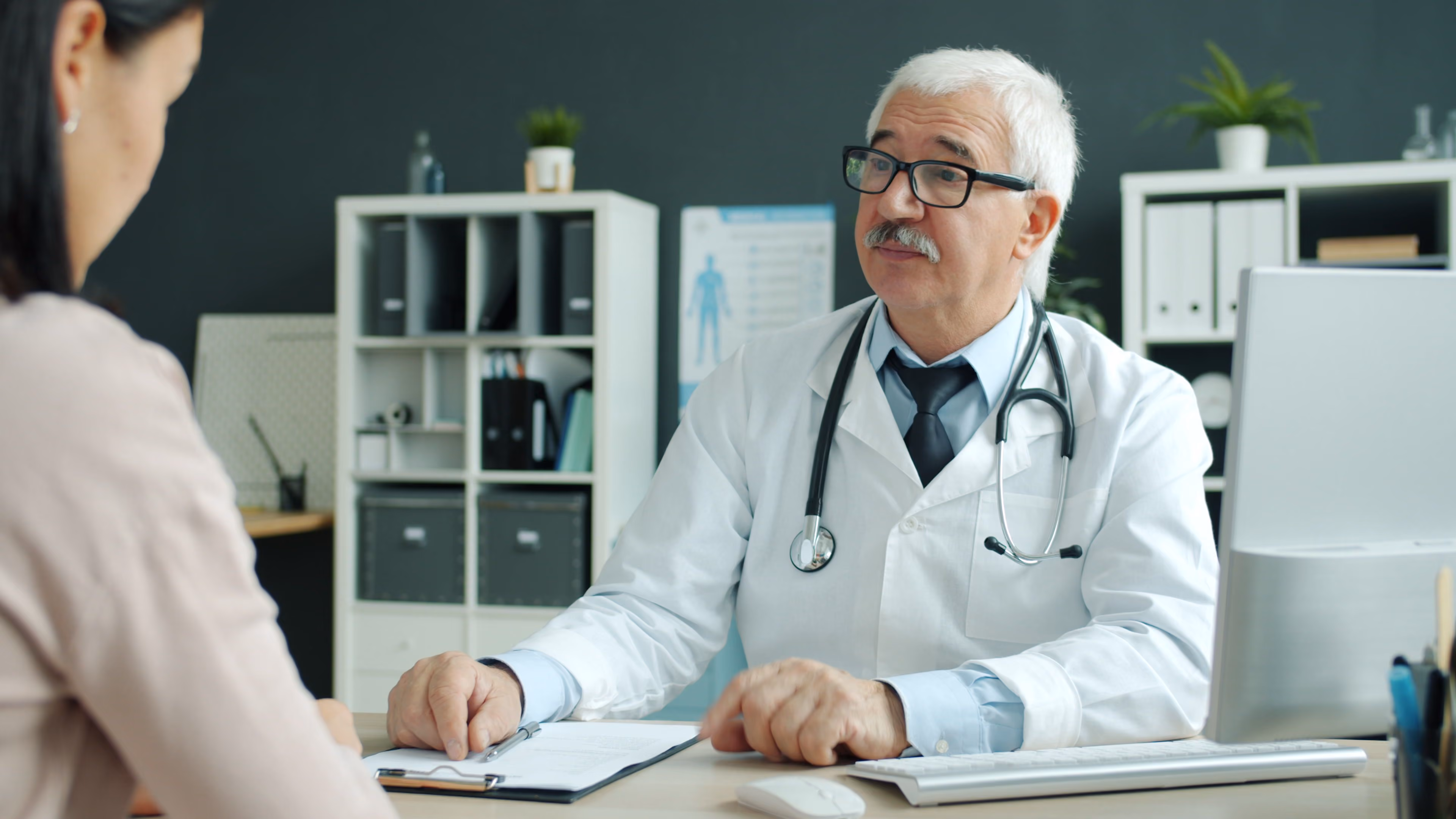 An older male doctor with white hair, glasses, and a stethoscope around his neck is sitting at a desk, talking to a female patient in an office with shelves and a computer.