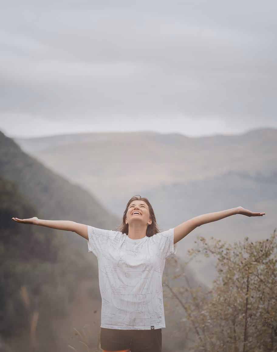 A person standing outdoors with arms outstretched and head tilted back, smiling, against a backdrop of misty mountains and trees.