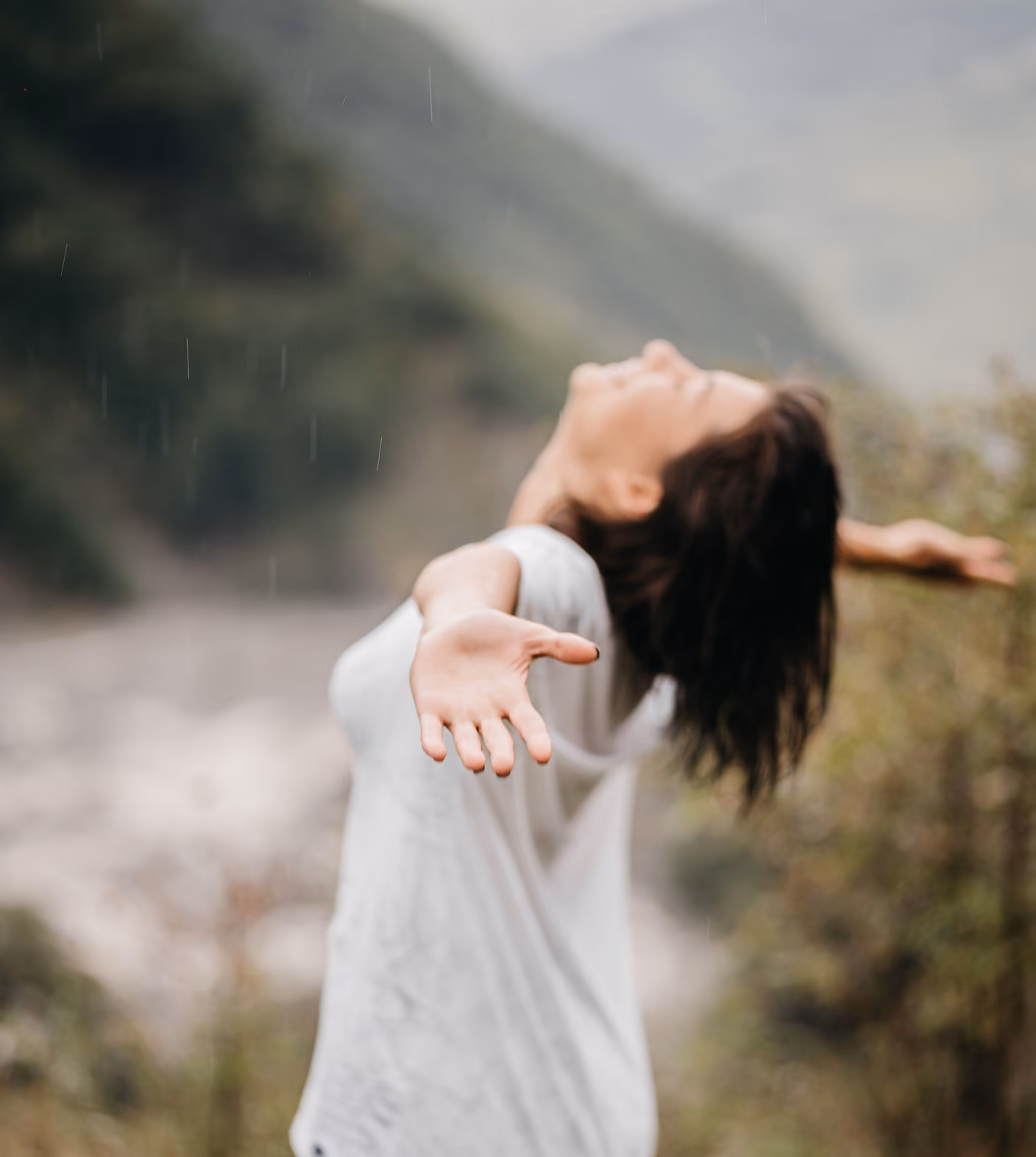 Person with outstretched arms and head tilted back enjoying a light rain outdoors, surrounded by blurred greenery and hills.