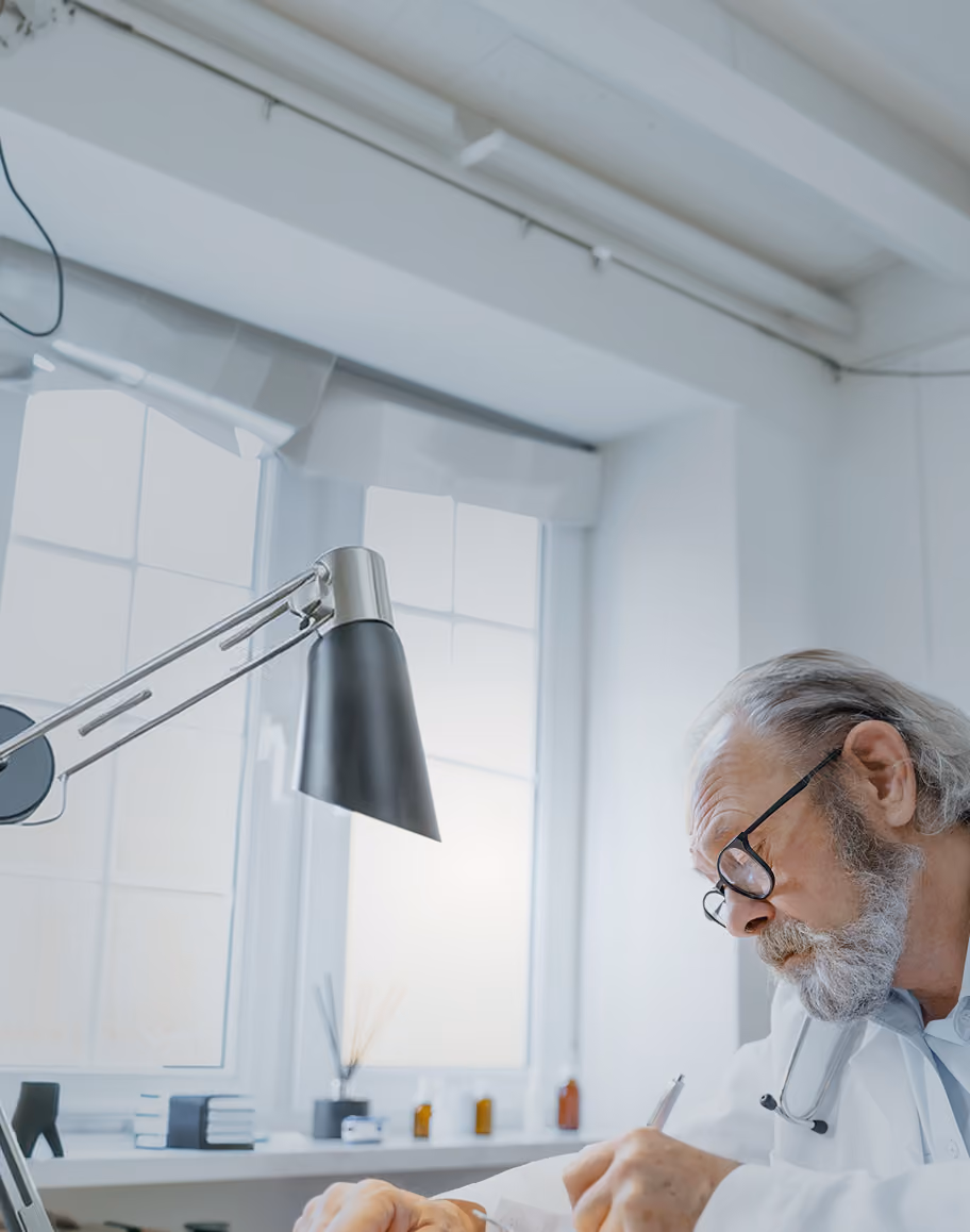 Older male doctor with glasses and grey hair attentively writing notes, wearing a stethoscope around his neck, in a bright office with large windows and a modern desk lamp.