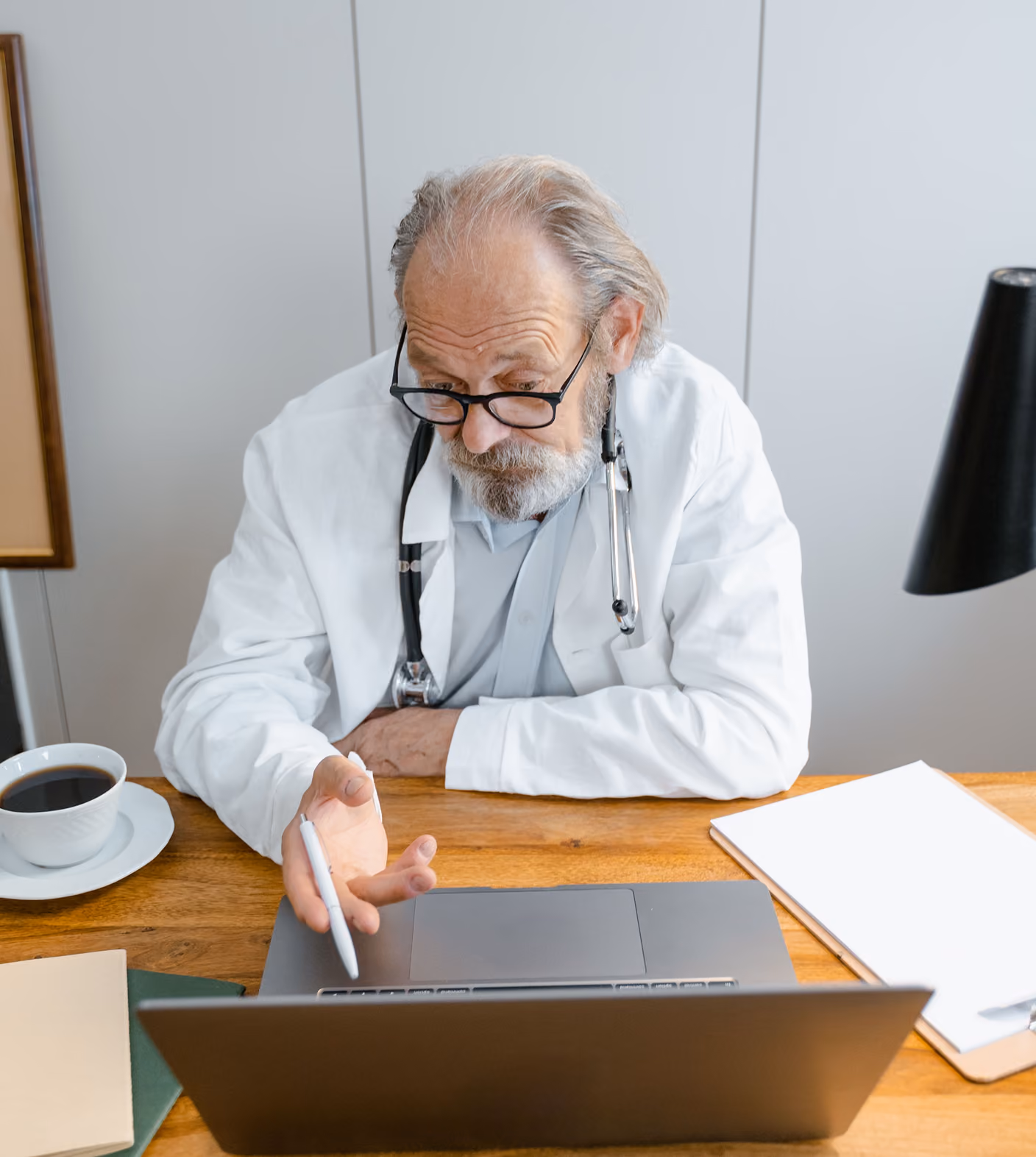 Older male doctor in a white coat and glasses sitting at a wooden desk, pointing with a pen at a laptop screen, with a stethoscope around his neck, a cup of coffee and folders on the desk.