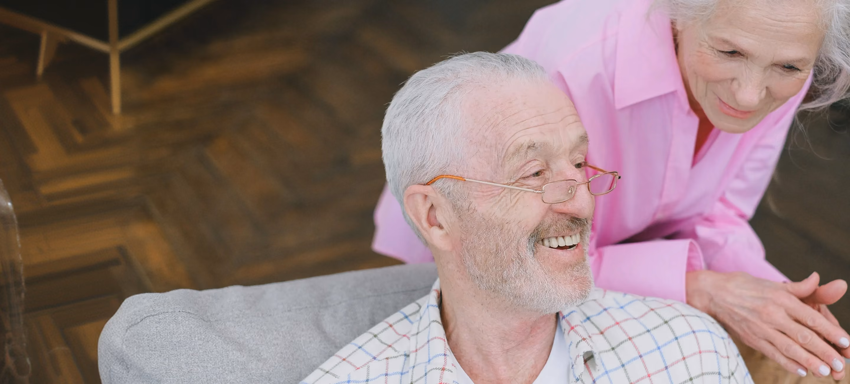 Smiling elderly man with gray hair and glasses sitting on a gray couch, wearing a white shirt with a colorful checkered pattern; an elderly woman with gray hair and pink blouse leans over him with a gentle smile.