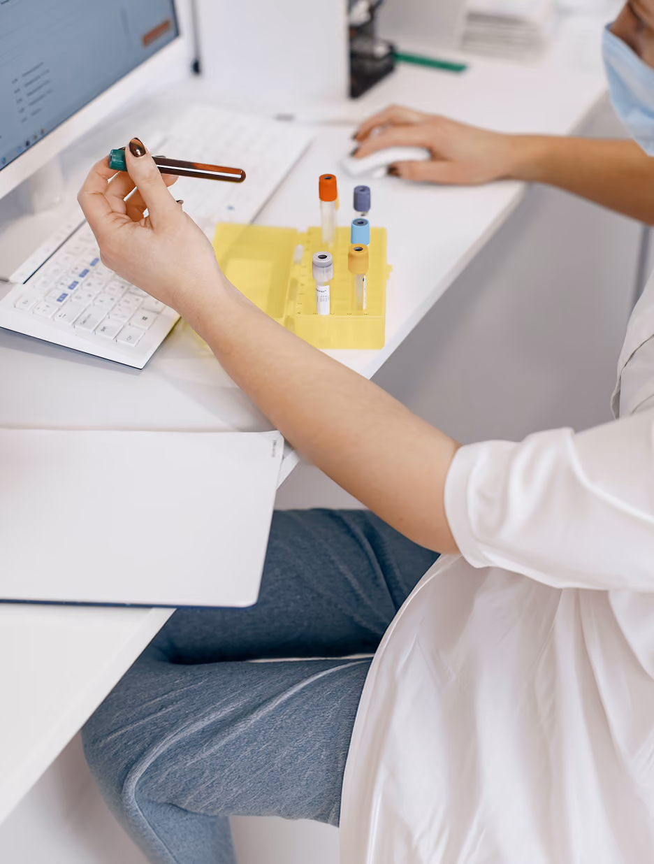 Medical professional wearing a mask and white coat holding a blood sample tube while working at a computer with multiple sample tubes in a yellow holder on the desk.