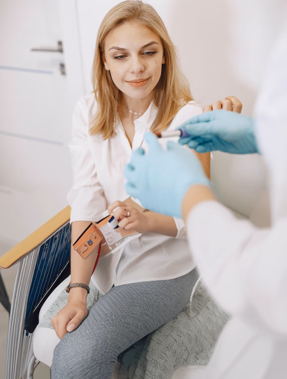 A woman sitting on a chair with a blood pressure cuff on her arm, looking at a healthcare professional wearing blue gloves who is holding a medical device.