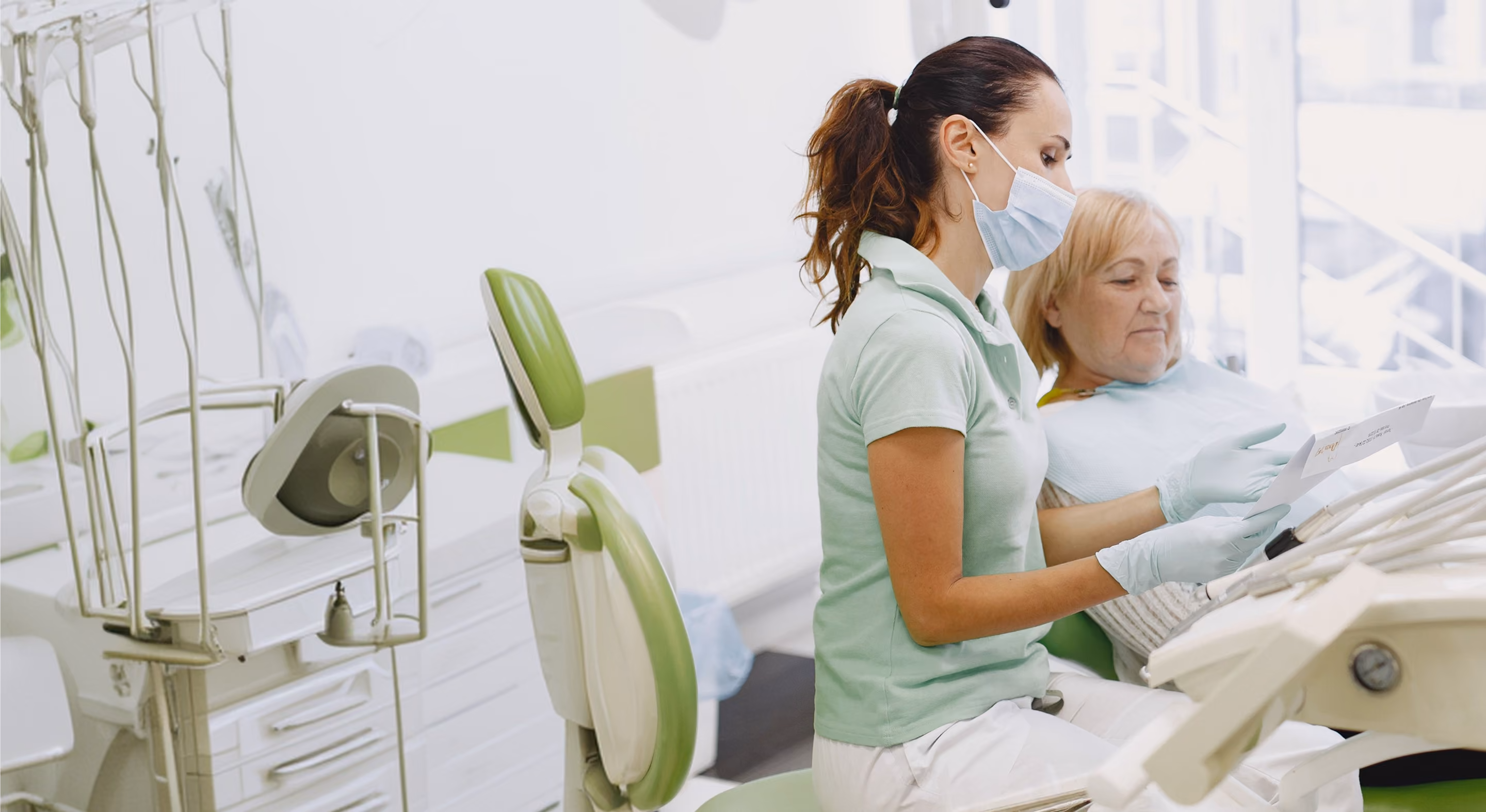 A dentist wearing a mask and gloves shows a document to an older female patient sitting in a dental chair in a bright dental clinic.