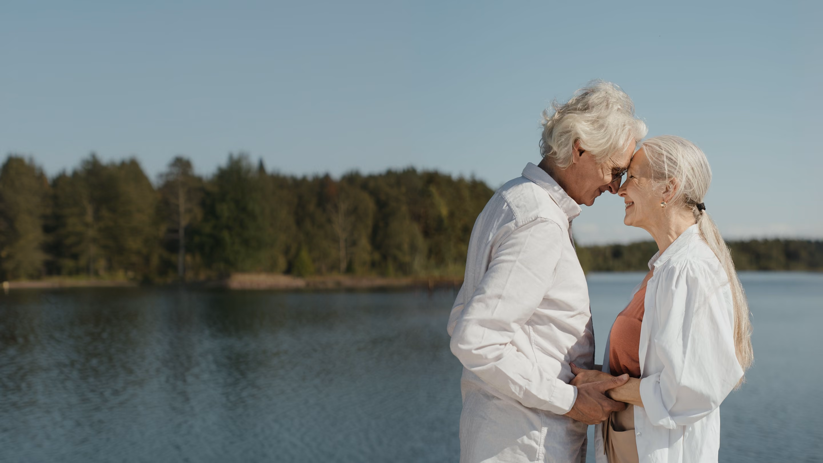 Elderly couple standing close together by a lake with a forest and clear blue sky in the background, holding hands and smiling at each other.