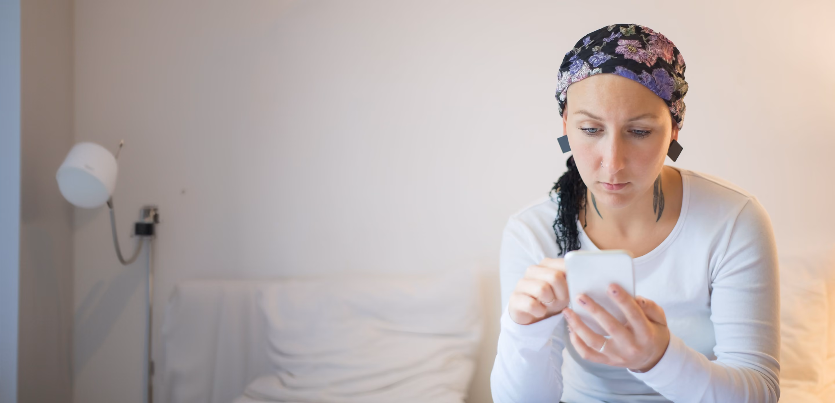 Woman wearing a floral headscarf and white long-sleeve shirt, seated on a white couch, intently looking at her smartphone in a softly lit room.