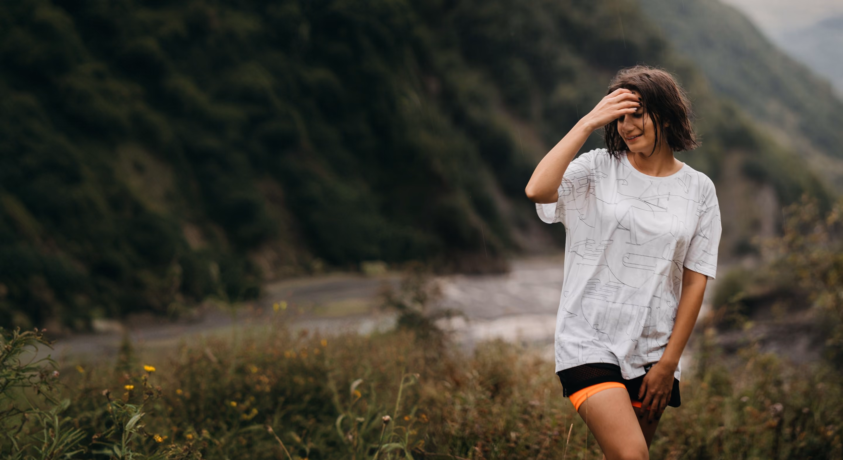 Woman standing outdoors in front of a blurred river and forested hills, wearing a white t-shirt and orange shorts, with one hand on her head and the other on her thigh.