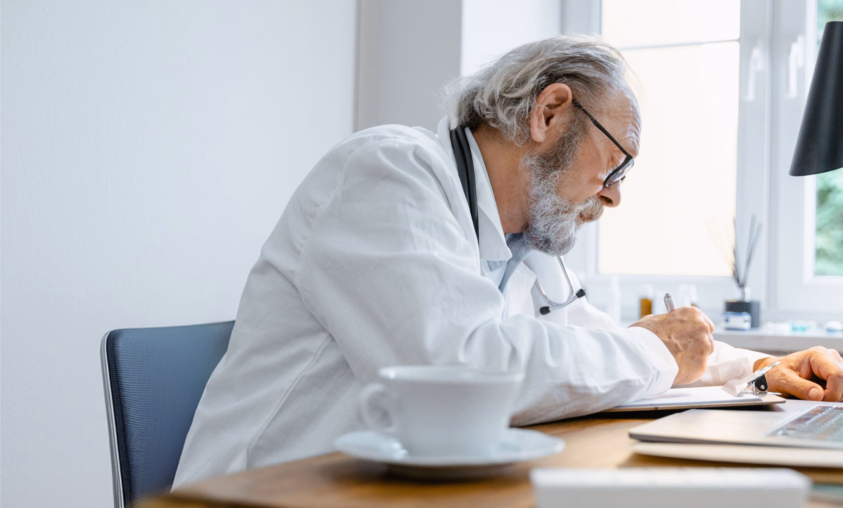 Male doctor with white hair and beard wearing a white coat and glasses, sitting at a desk near a window, writing on a clipboard with a stethoscope around his neck, with a white coffee cup and a laptop on the desk.