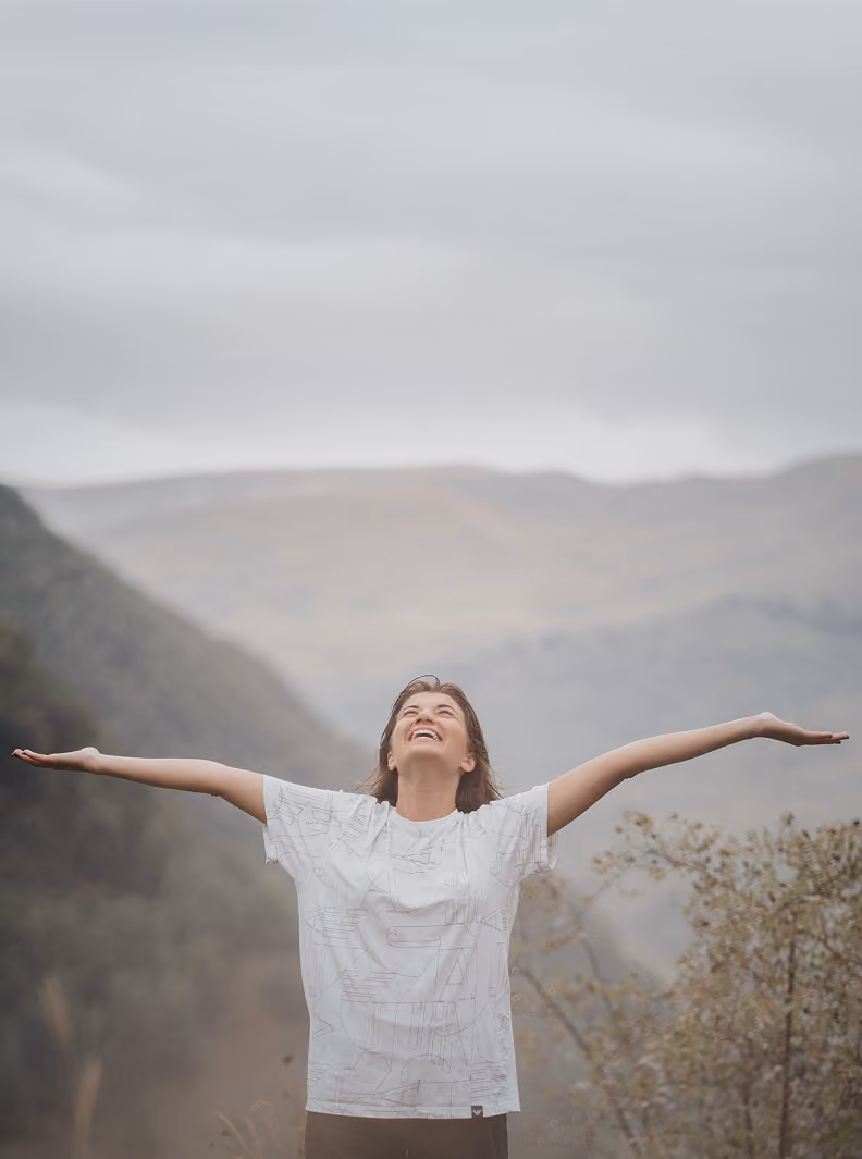 Young woman standing outdoors with arms outstretched and head tilted back, smiling, against a backdrop of misty mountains and overcast sky.