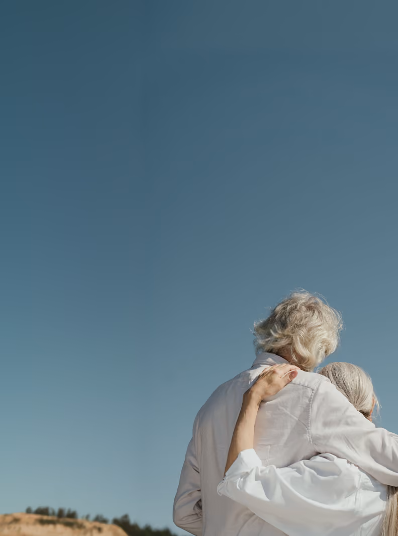 Senior couple embracing outdoors against a clear blue sky, seen from behind.