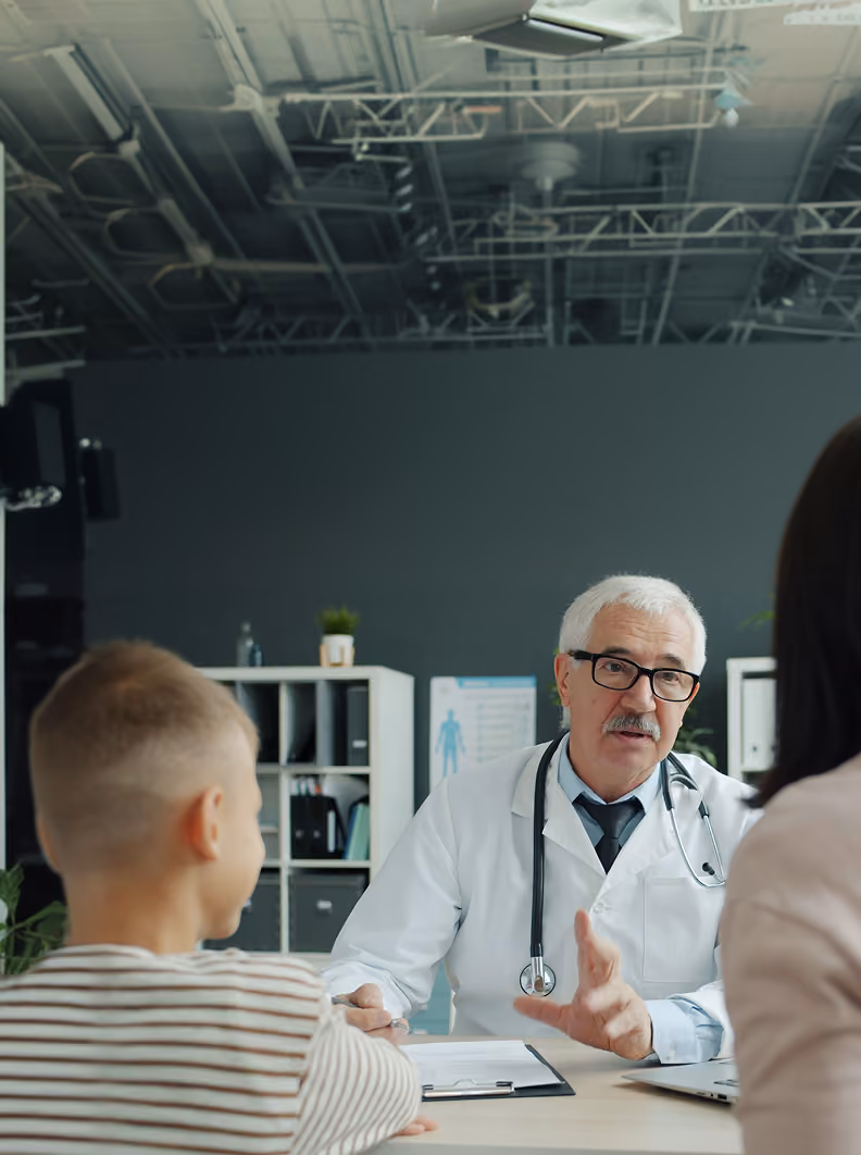 Elderly male doctor with white hair, glasses, and a stethoscope around his neck talking to a young boy and woman in a modern medical office.