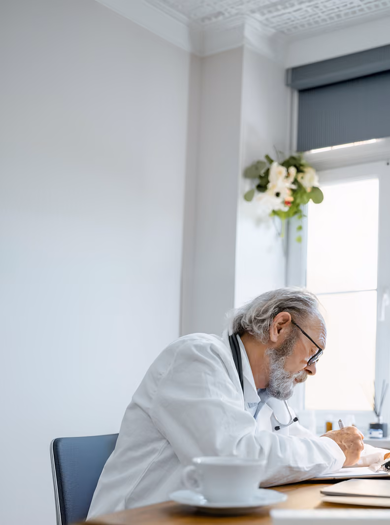 Older male doctor with gray hair and beard wearing glasses and a white coat, writing on a clipboard at a desk with a white cup in the foreground in a bright room with a window and a flower decoration on the wall.