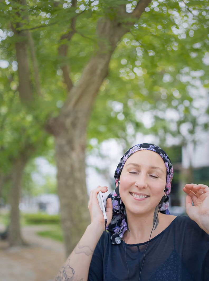 A smiling woman wearing a floral headscarf and black top listens to music with earphones outdoors in a leafy park.