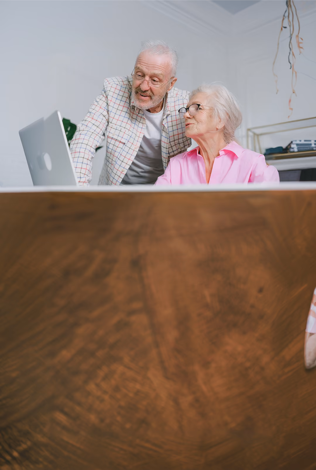 An elderly man and woman wearing glasses looking at a laptop screen together, with the man standing and the woman seated at a wooden table.