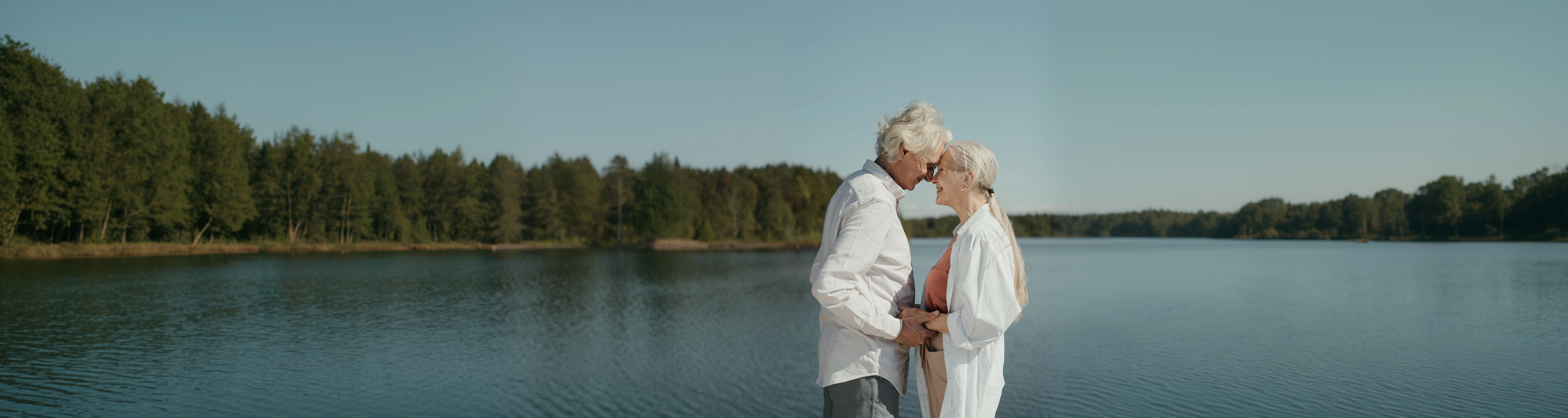 An elderly couple stands close together by a calm lake, holding hands and smiling with their foreheads touching, surrounded by trees under a clear sky.