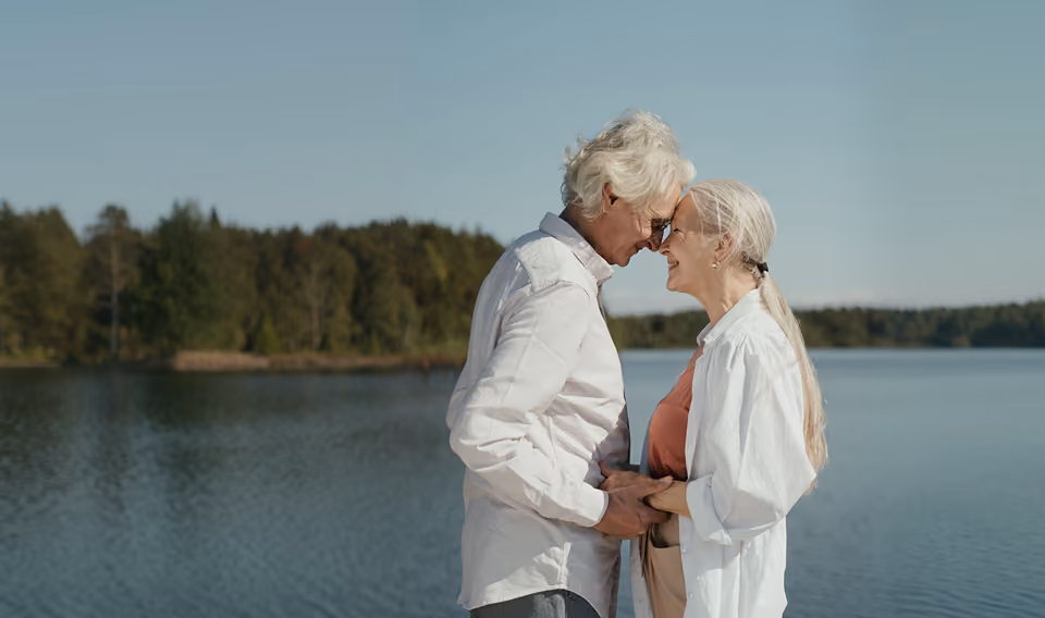 An elderly couple stands close together by a calm lake, holding hands and smiling with their foreheads touching, surrounded by trees under a clear sky.