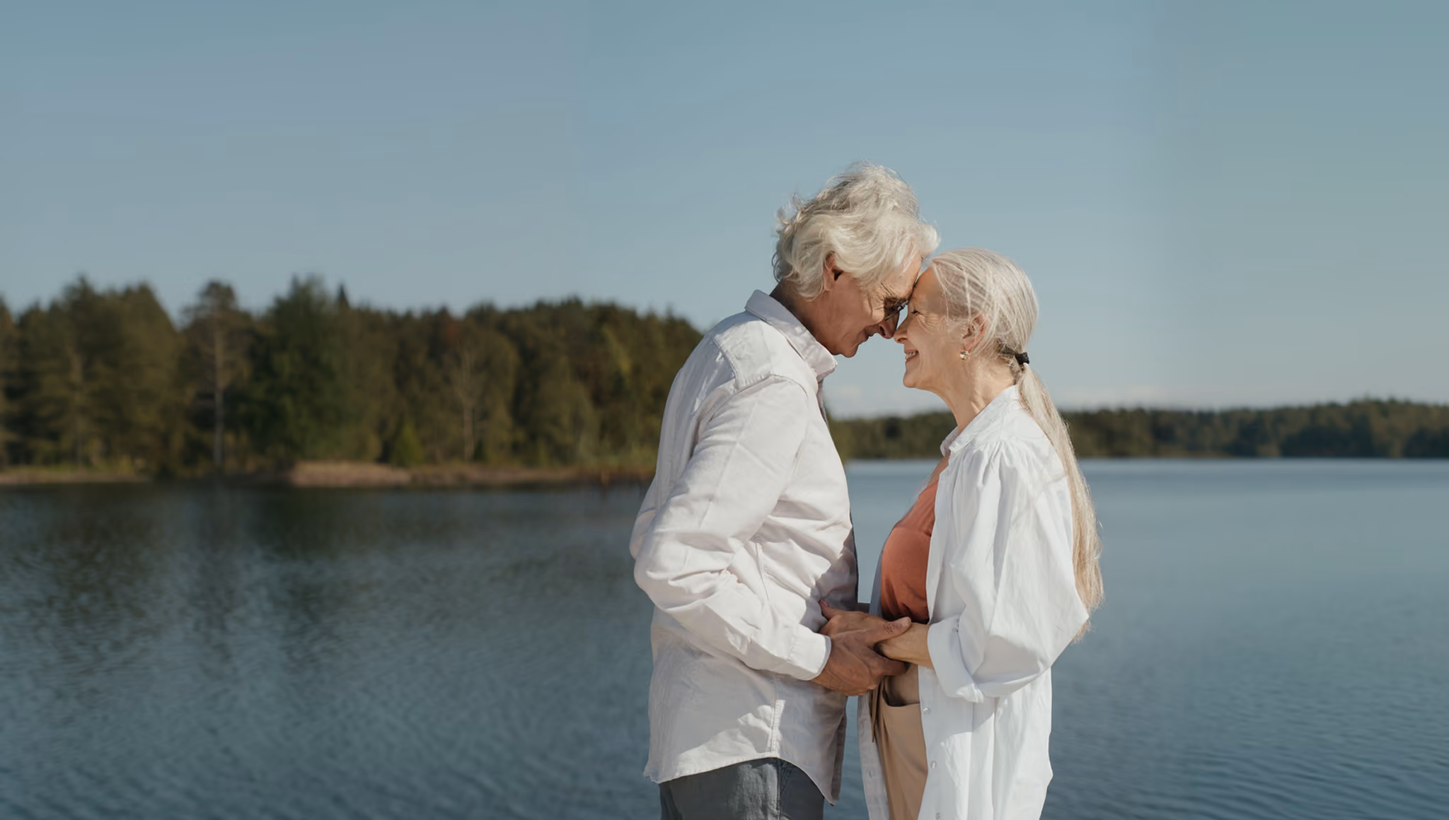An elderly couple stands close together by a calm lake, holding hands and smiling with their foreheads touching, surrounded by trees under a clear sky.